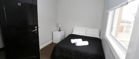 A hotel room with a black door marked with the number 12, a bed with a black bedspread and two white towels, a small bedside table with a lamp, and a window with a view of the outside.