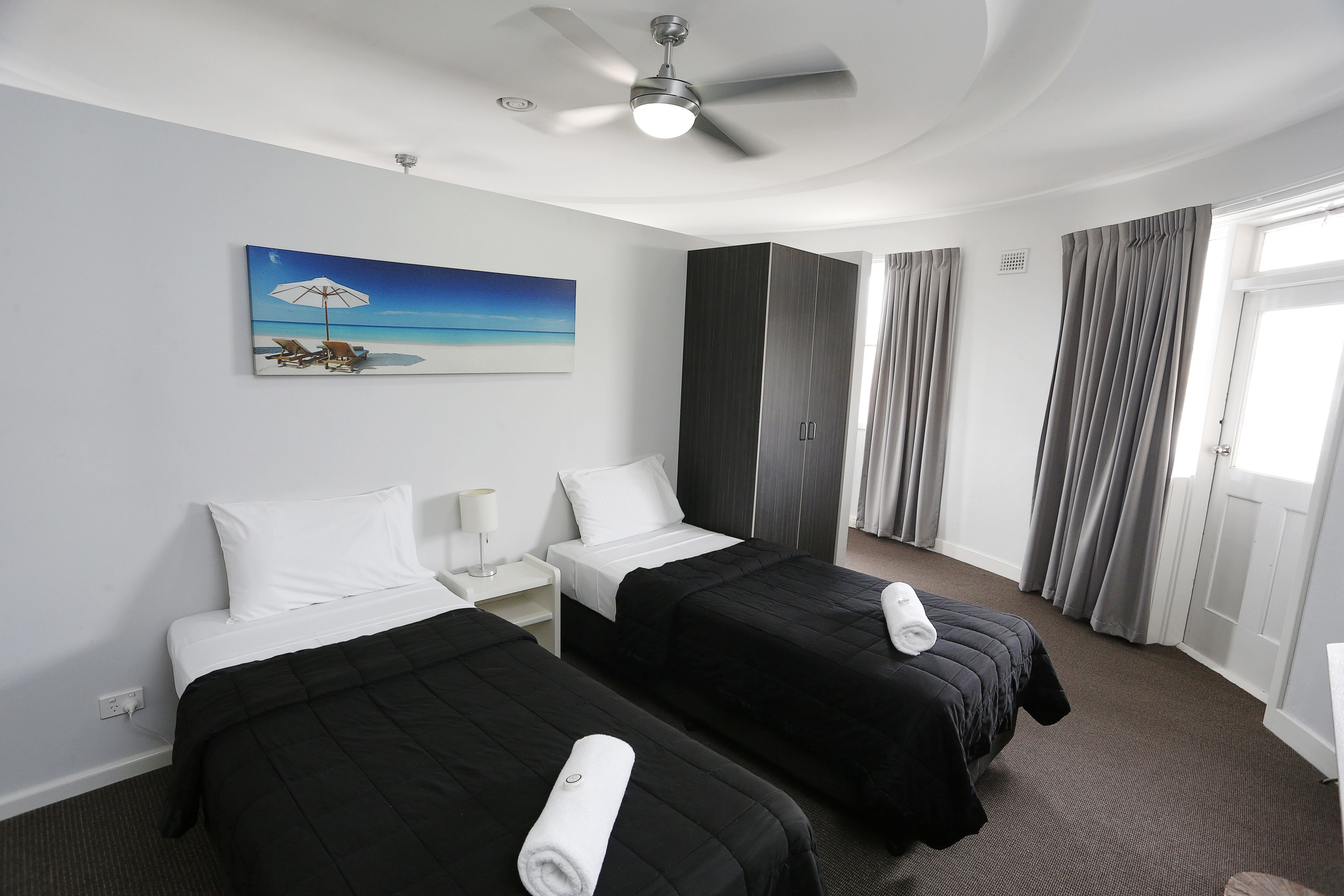 A hotel room with two beds, a ceiling fan, a painting of a beach scene, and a large window with curtains.