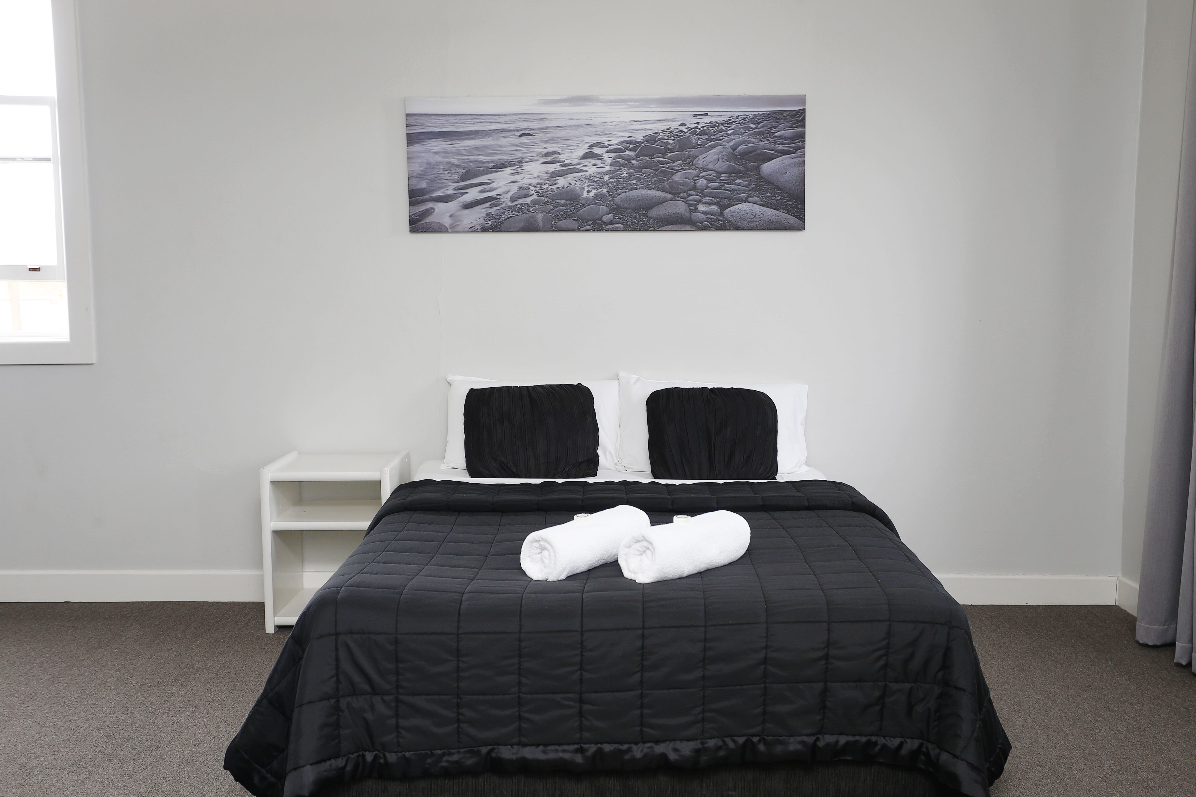 A neatly made bed with a black quilt, two pillows, and rolled towels. A white nightstand is beside the bed. A large black and white photograph of a rocky beach is on the wall.