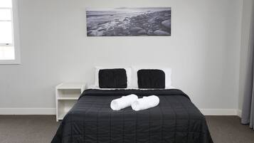A neatly made bed with a black quilt, two pillows, and rolled towels. A white nightstand is beside the bed. A large black and white photograph of a rocky beach is on the wall.