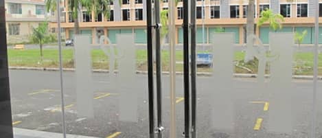 Vista a través de una puerta de cristal con un tapete rojo, palmeras y un edificio con varias ventanas al fondo.