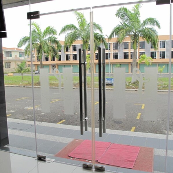 A view through a glass door with a red mat, palm trees, and a building with multiple windows in the background. - Labuk Hotel (Sandakan)