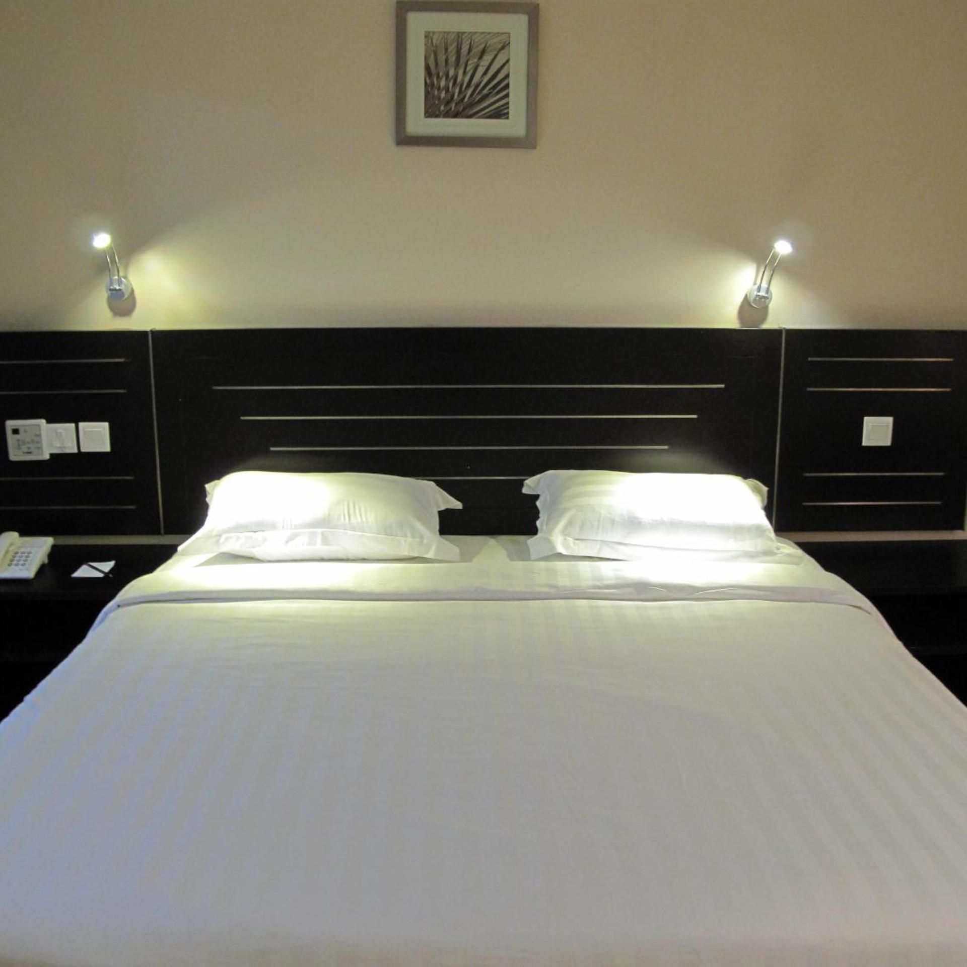 A hotel room with a bed, two bedside tables, a nightstand, a framed picture on the wall, and two wall-mounted lamps.