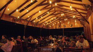 Een eethoek inpandig, met houten balken en hangende lampen. Tafels zijn gedekt met stoelen, en mensen zitten.