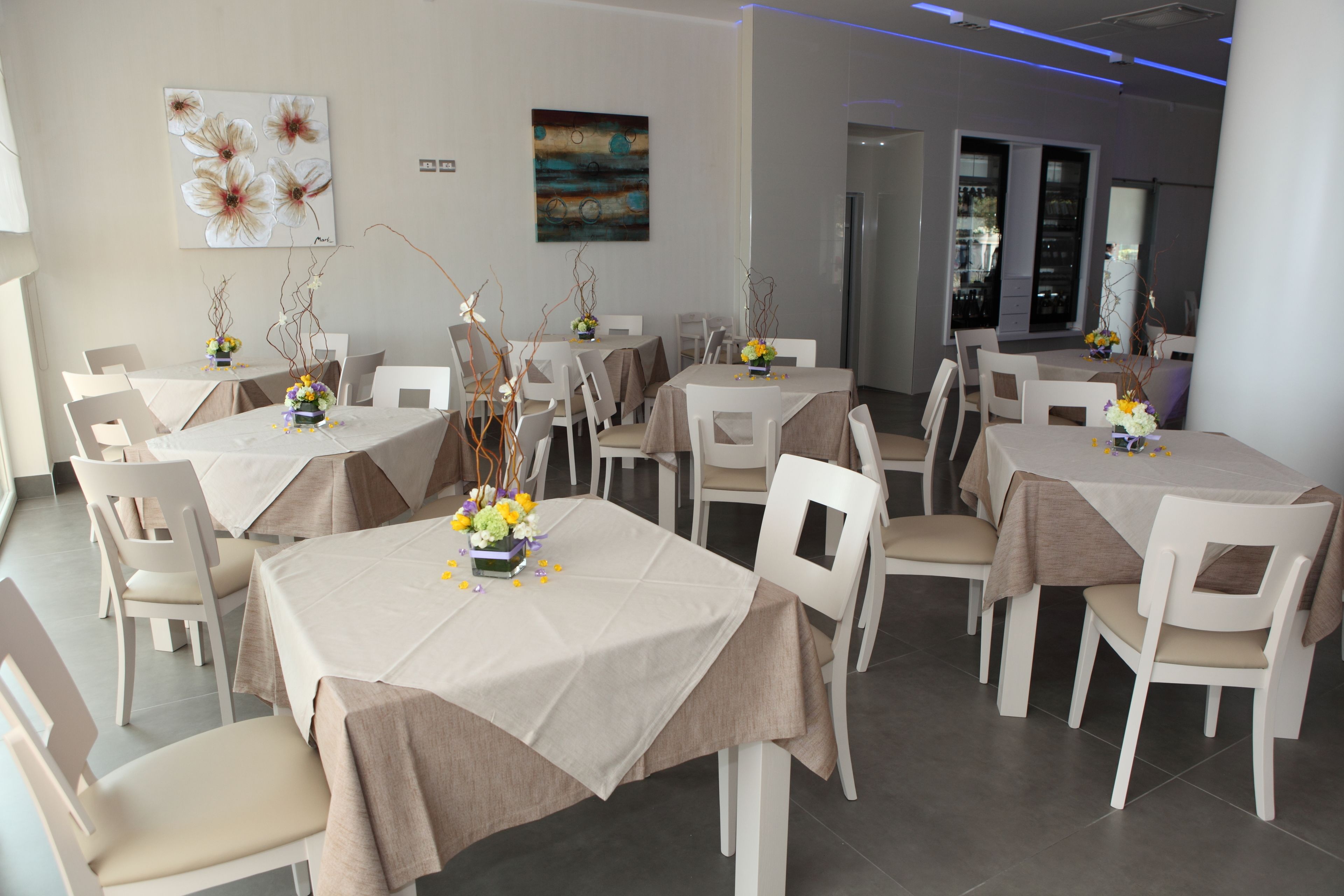 Zona pranzo con diversi tavoli e sedie, ognuno con tovaglia e centrotavola floreale.