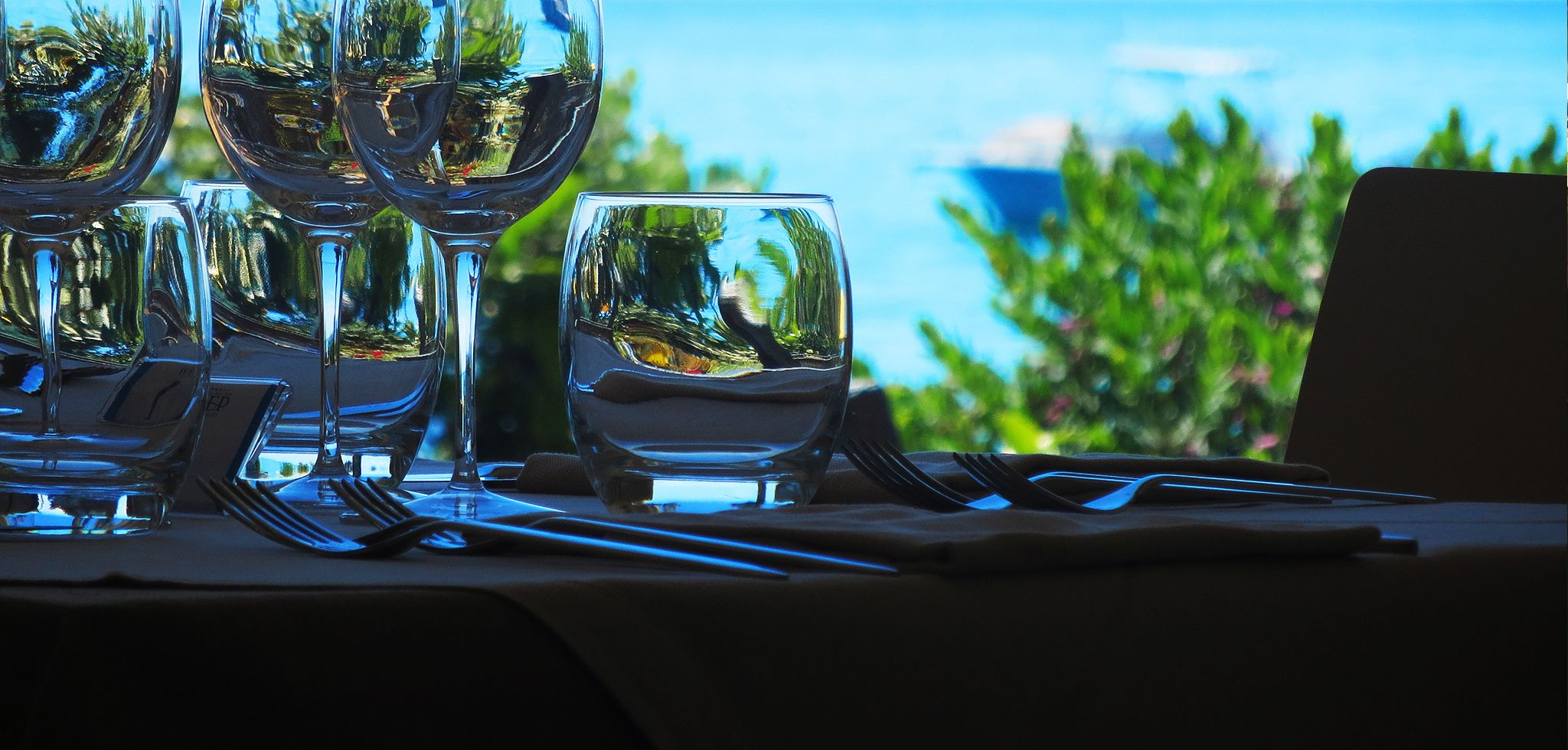 Un tavolo apparecchiato con bicchieri da vino e un bicchiere d'acqua, con vista su spazi verdi e il mare sullo sfondo.