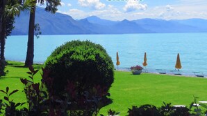 A serene lakeside view with a well-maintained lawn, a decorative bush, and a row of sun umbrellas along the water's edge. - Hotel Eden Park Cilento (Ispani)