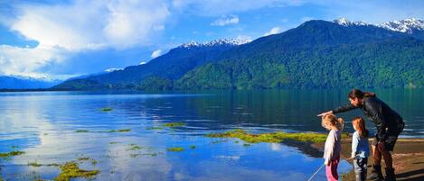 Un lago con montañas al fondo, cielo despejado y una persona señalando algo en el agua.