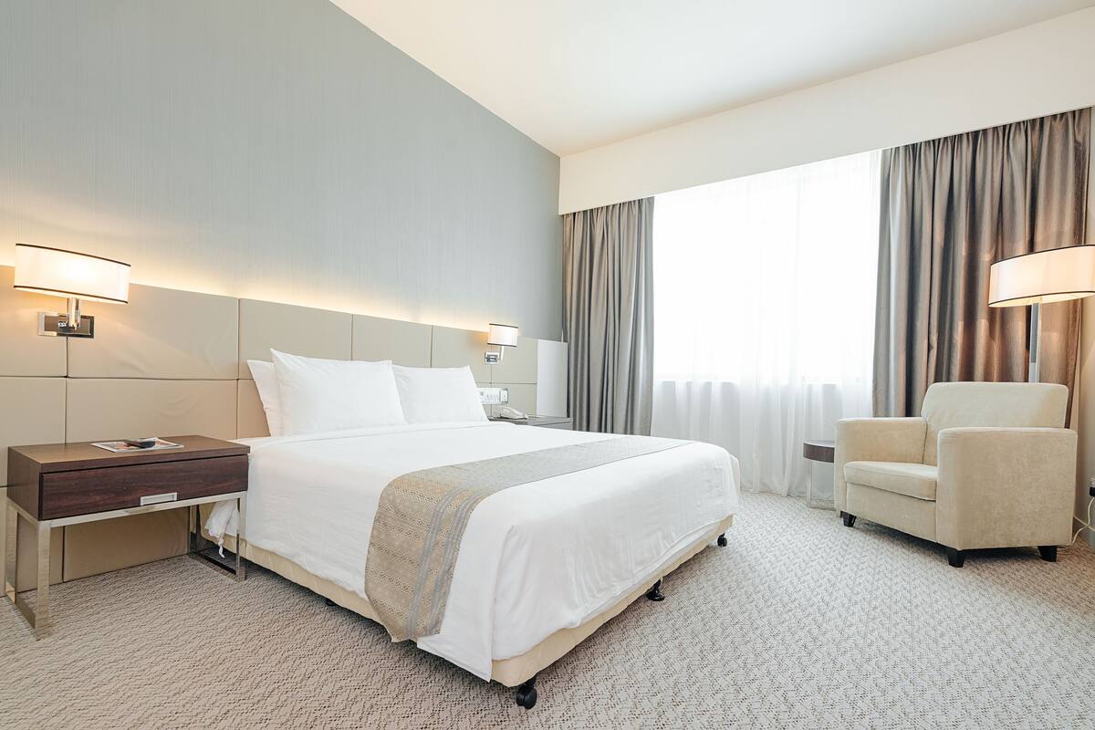 Quarto de hotel com cama grande, mesas de cabeceira, uma cadeira e janela com cortinas.