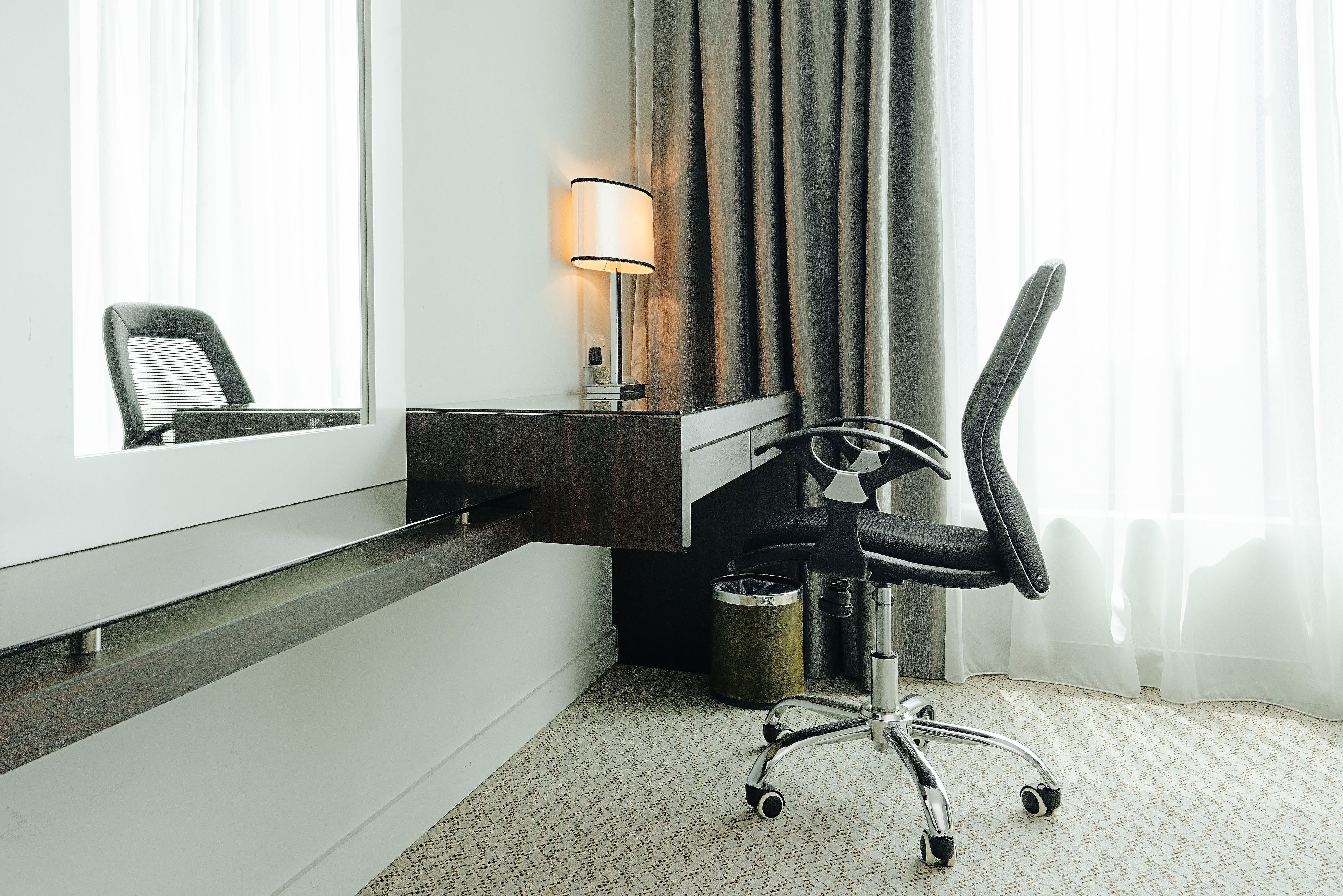 Una habitación de hotel con escritorio, silla, lámpara y un ventanal con cortinas.