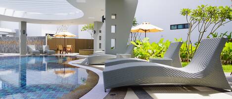 Un área de piscina moderna con sillas de descanso y sombrillas, rodeada de vegetación y un edificio contemporáneo.