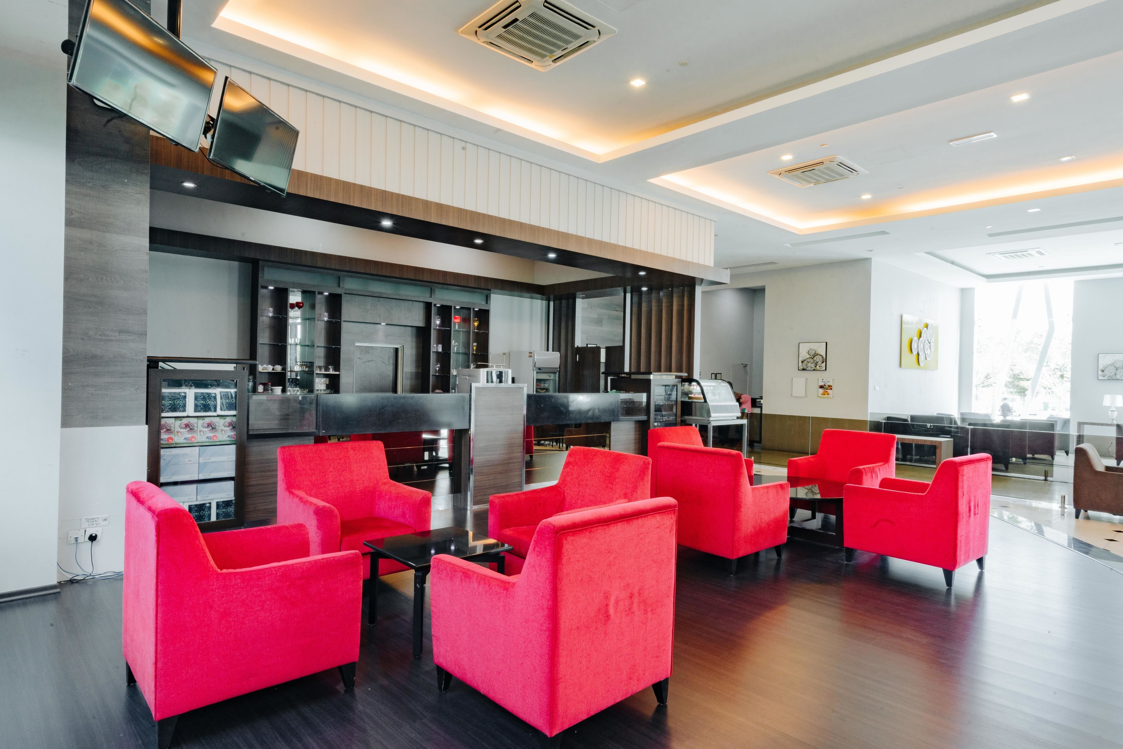 A modern hotel lobby with red chairs, a kitchen area, and a flat-screen TV mounted on the wall.