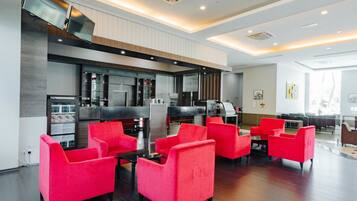 A modern hotel lobby with red chairs, a kitchen area, and a flat-screen TV mounted on the wall.