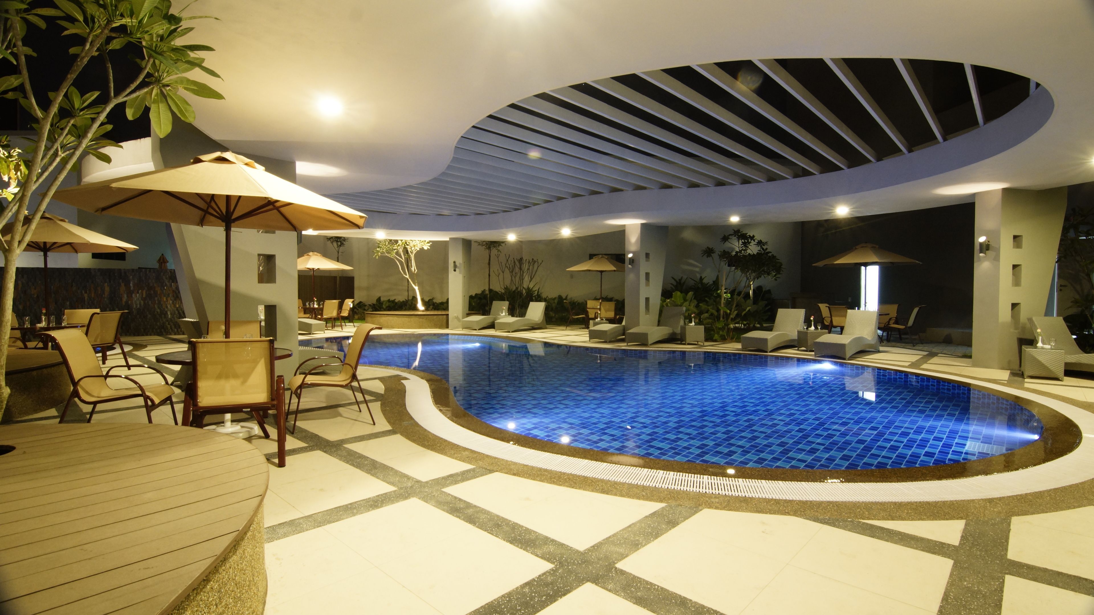 A modern indoor swimming pool with lounge chairs and umbrellas, surrounded by a tiled floor and a curved ceiling.