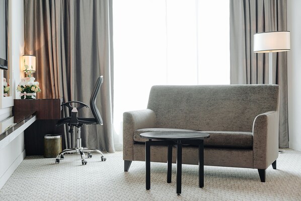 A modern hotel room with a grey sofa, a small round table, a black office chair, a lamp, and a trash can.