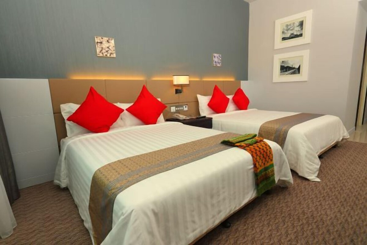 Habitación de hotel con dos camas, cada una con ropa de cama blanca y almohadas rojas. Hay luces fijadas a la pared y cuadros enmarcados.