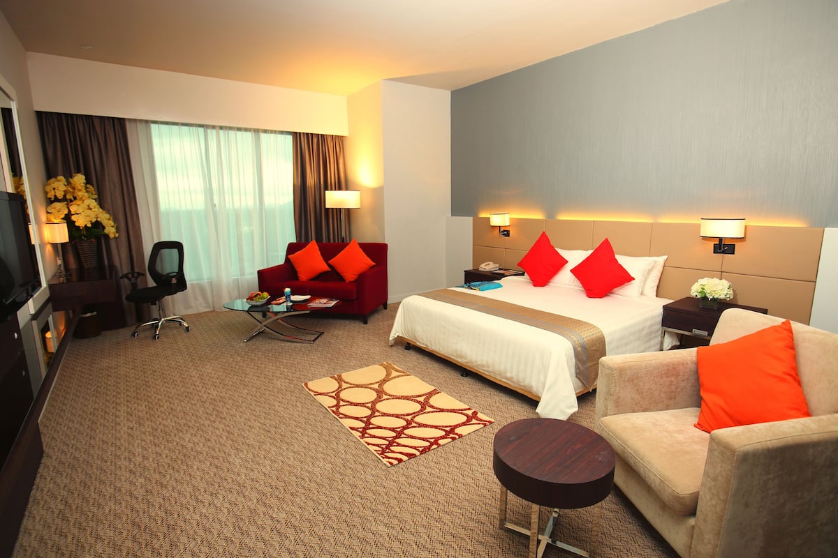 Quarto de hotel com cama, poltrona vermelha, sofá bege, escrivaninha e televisão.