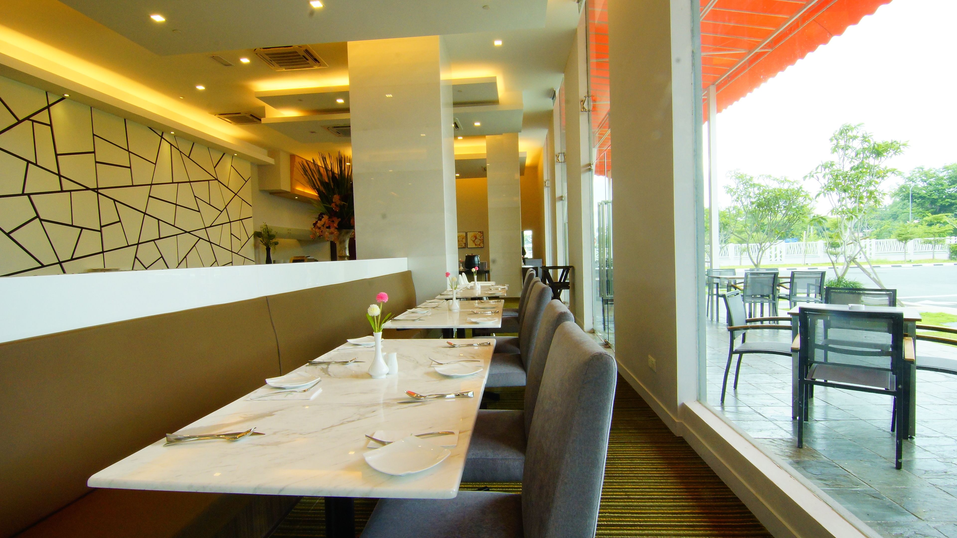 A restaurant with a modern interior, featuring a long dining area with tables set for two, a patterned wall, and a view of the outdoors.