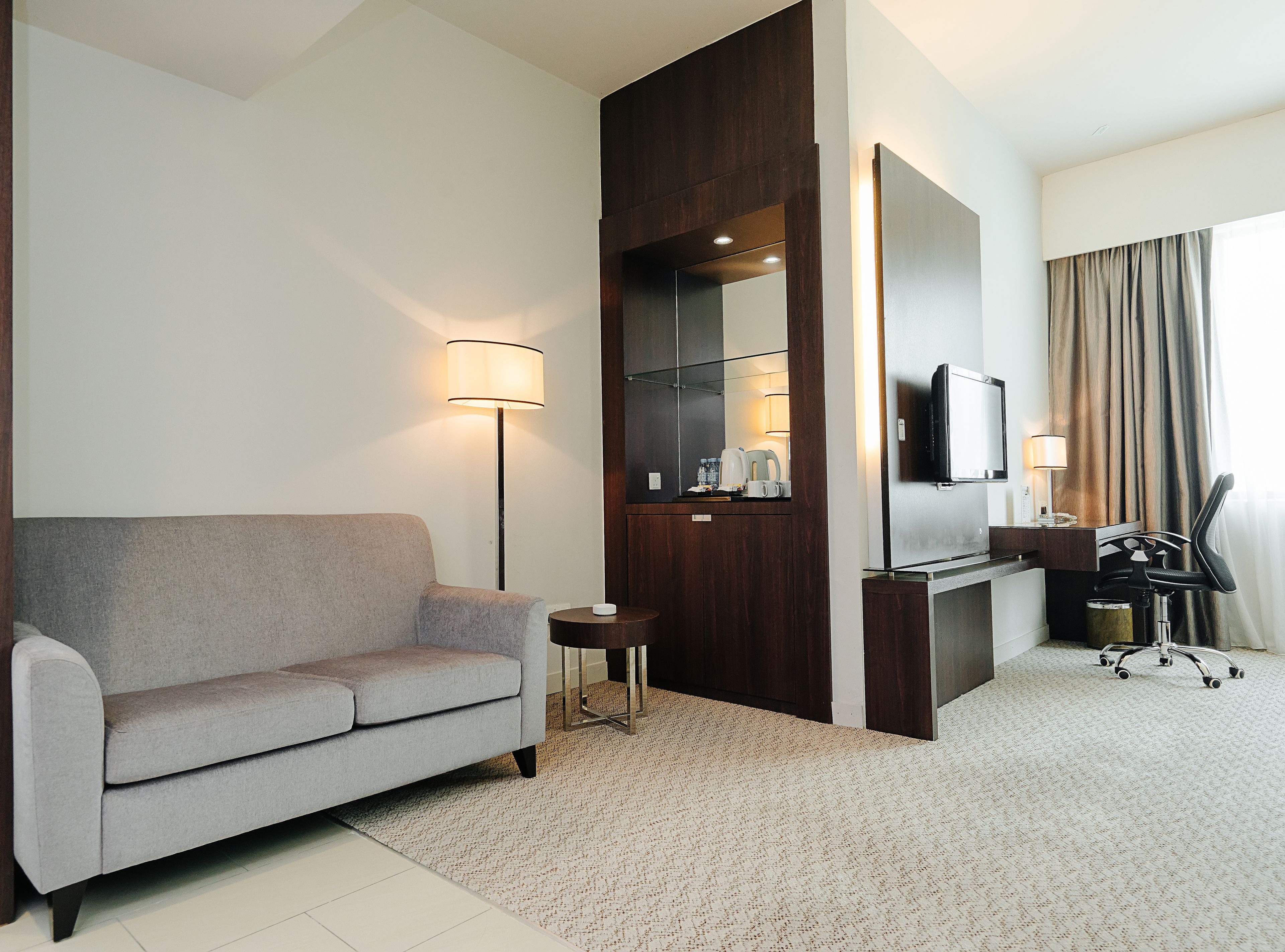 Een moderne hotelkamer met een grijze bank, een bureau met stoel, een flatscreen televisie, een lamp en een grote spiegel.