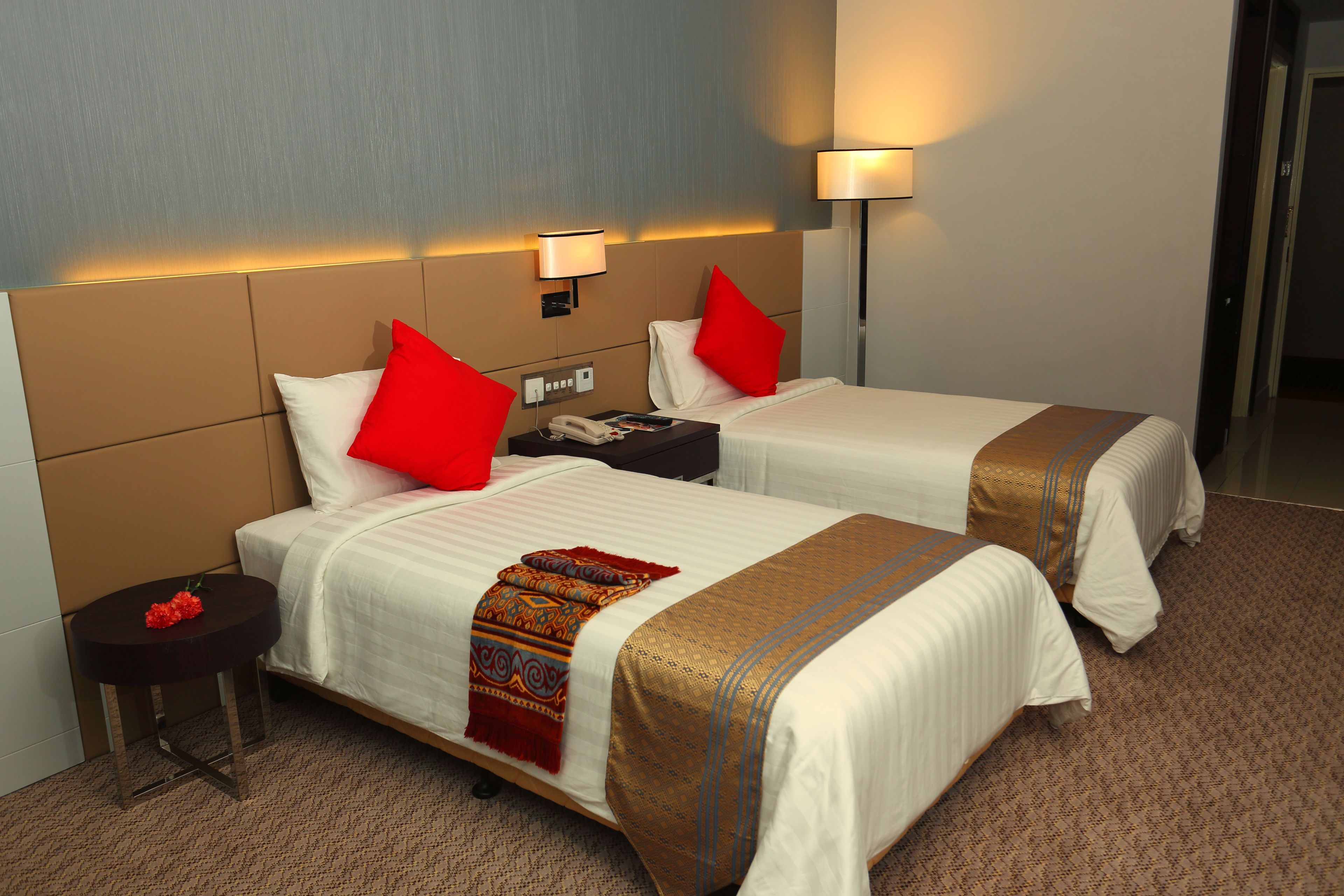 Habitación de hotel con dos camas, cada una con un cojín rojo, una mesita de noche con un teléfono y una lámpara, y suelo alfombrado.