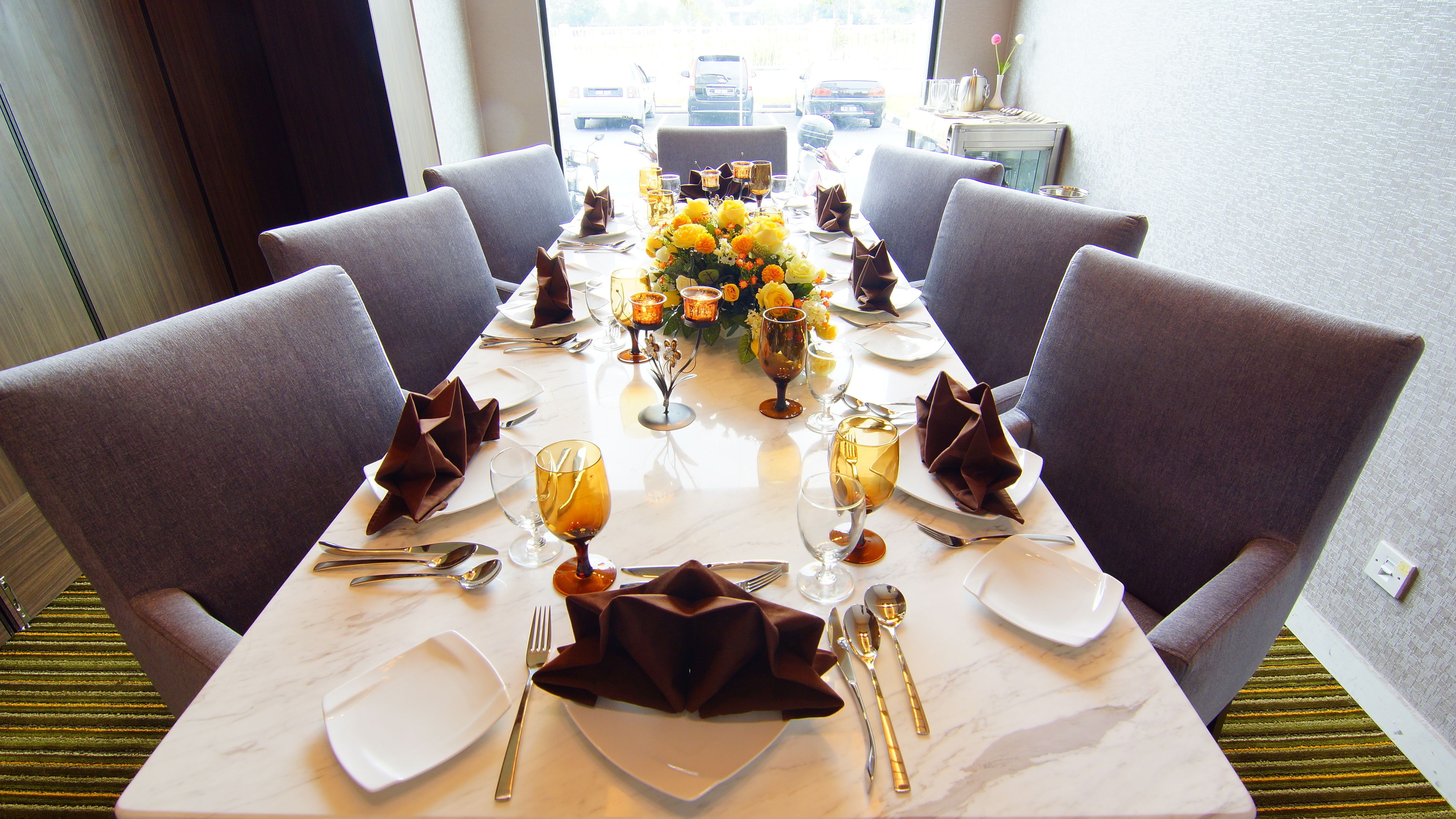 Mesa de comedor preparada para seis personas, con un florero de flores amarillas, velas y vista a coches estacionados afuera.
