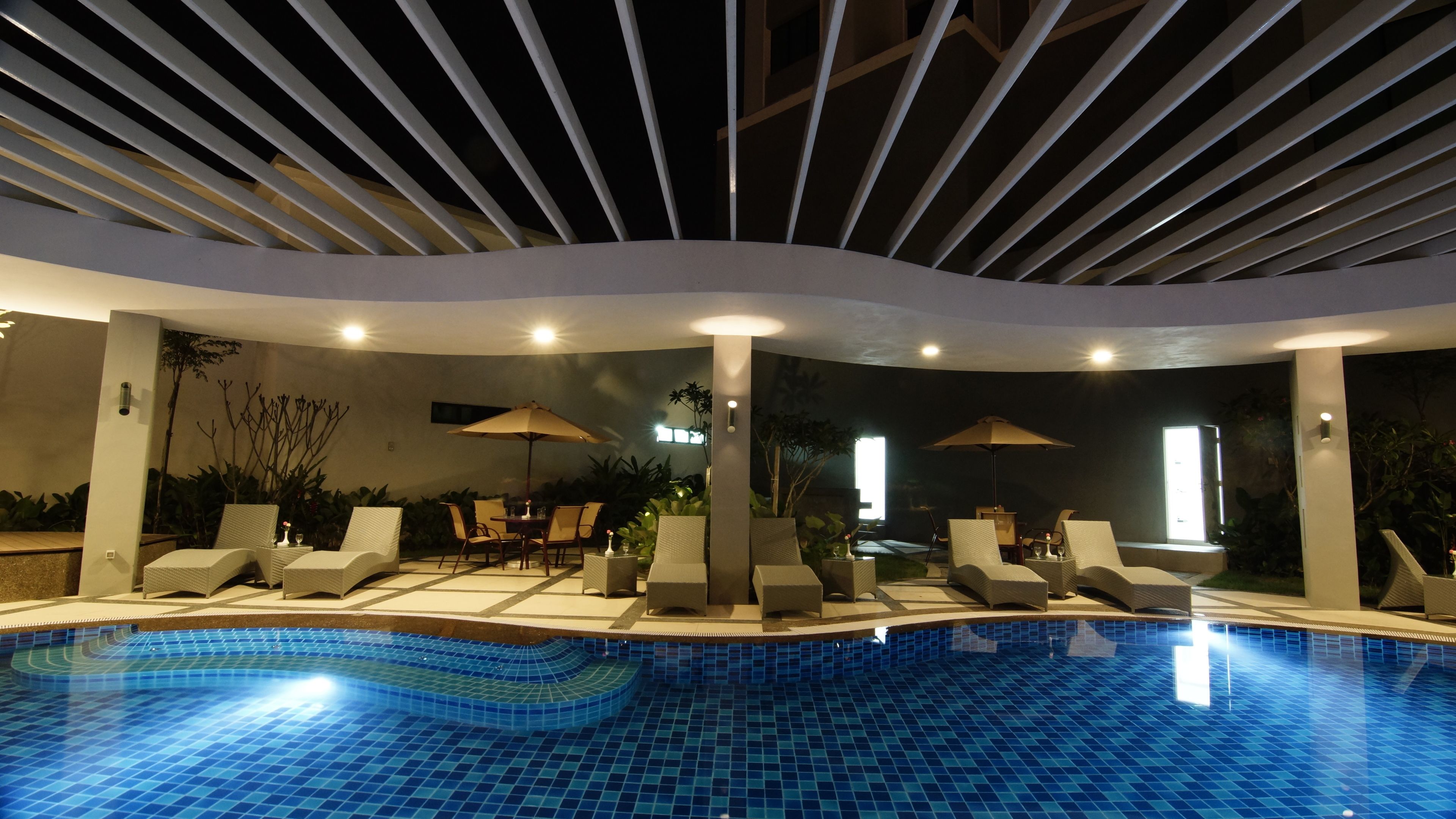 A pool area with lounge chairs and umbrellas, surrounded by walls with plants and a tiled floor.