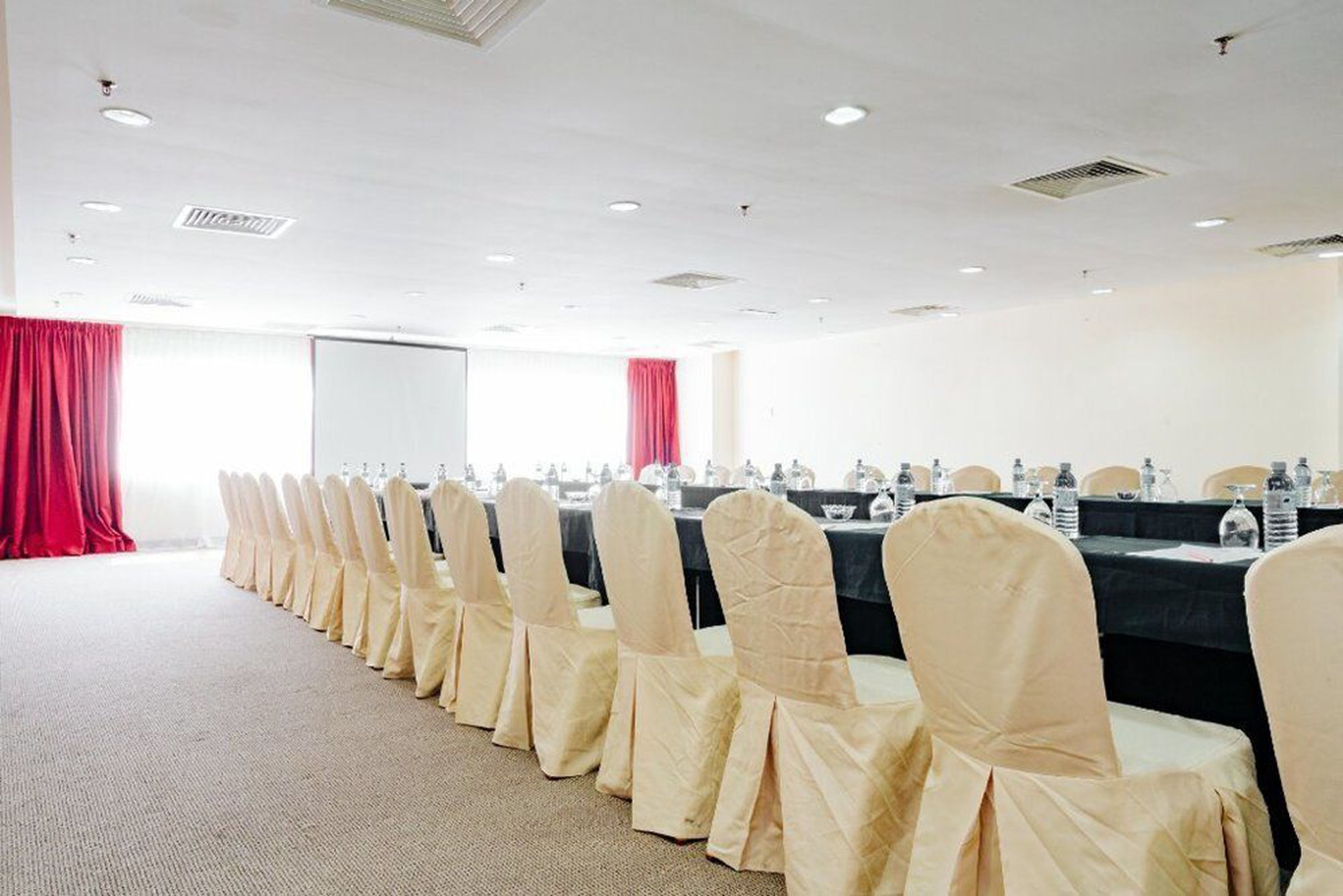 A conference room with rows of chairs covered in beige fabric, a projector screen, and tables with water bottles.