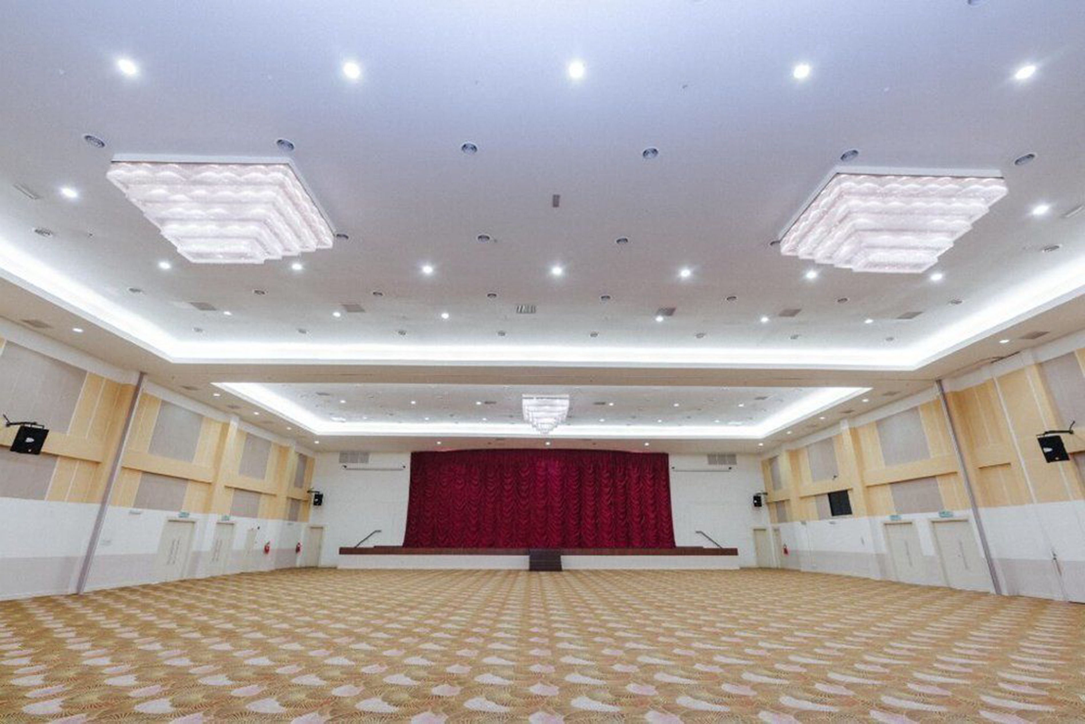 A large, well-lit conference room with a patterned carpet, a stage with a red curtain, and a modern ceiling design.