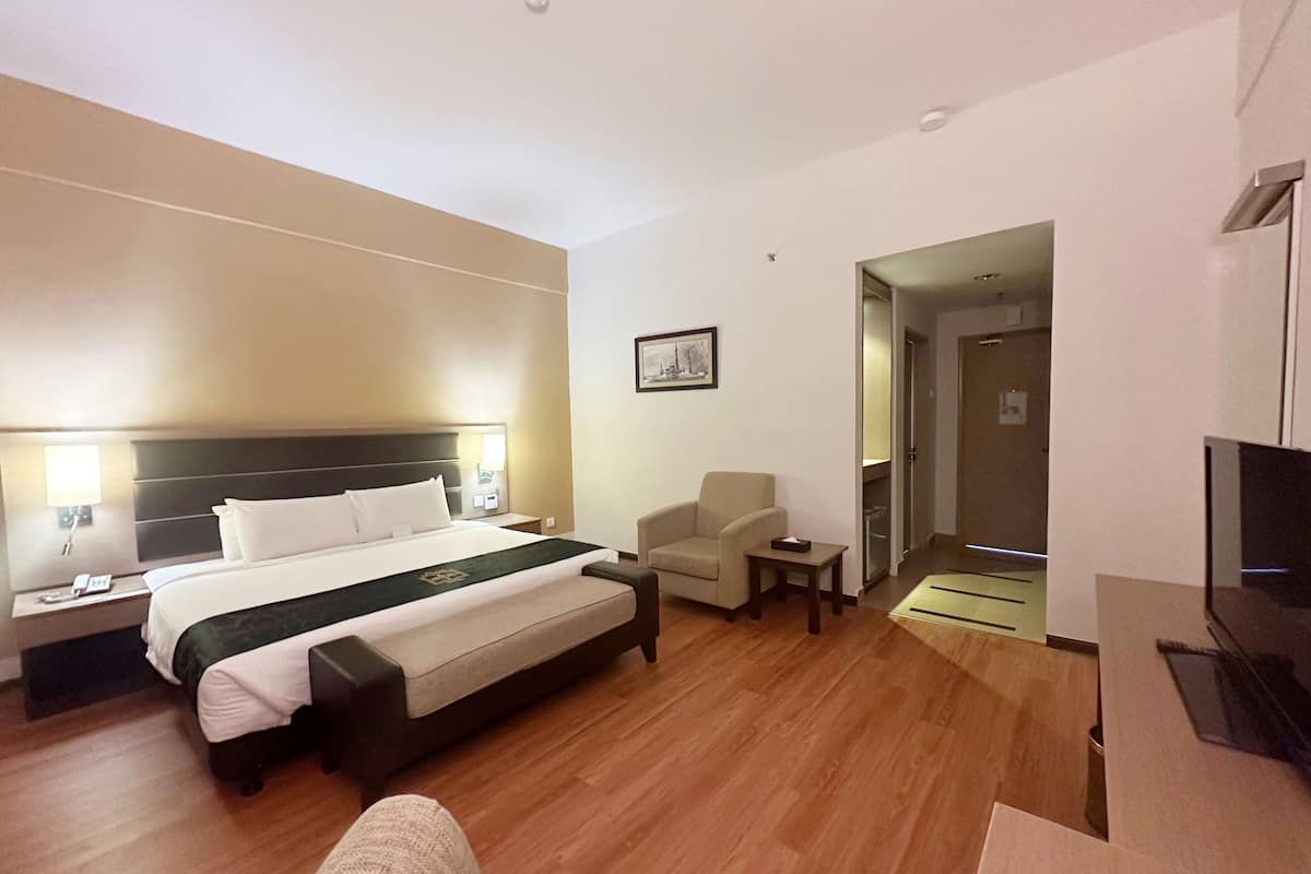 Une chambre d’hôtel avec un lit, une chaise, une petite table, un téléviseur et une porte donnant sur un couloir.