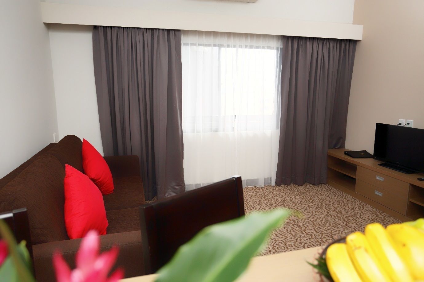 A hotel room with a brown sofa, red cushions, a television on a wooden stand, and a window with grey curtains.