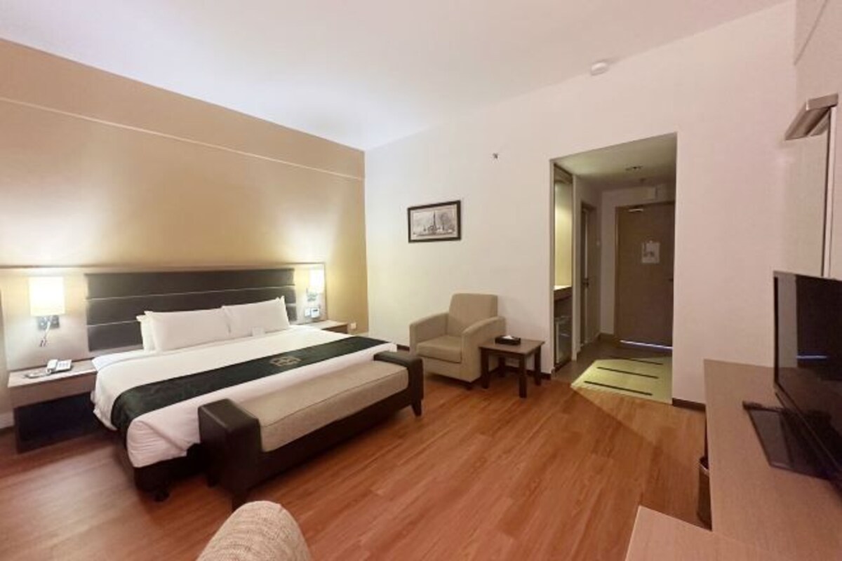 A hotel room with a bed, bedside table, chair, TV, and a door leading to another room.