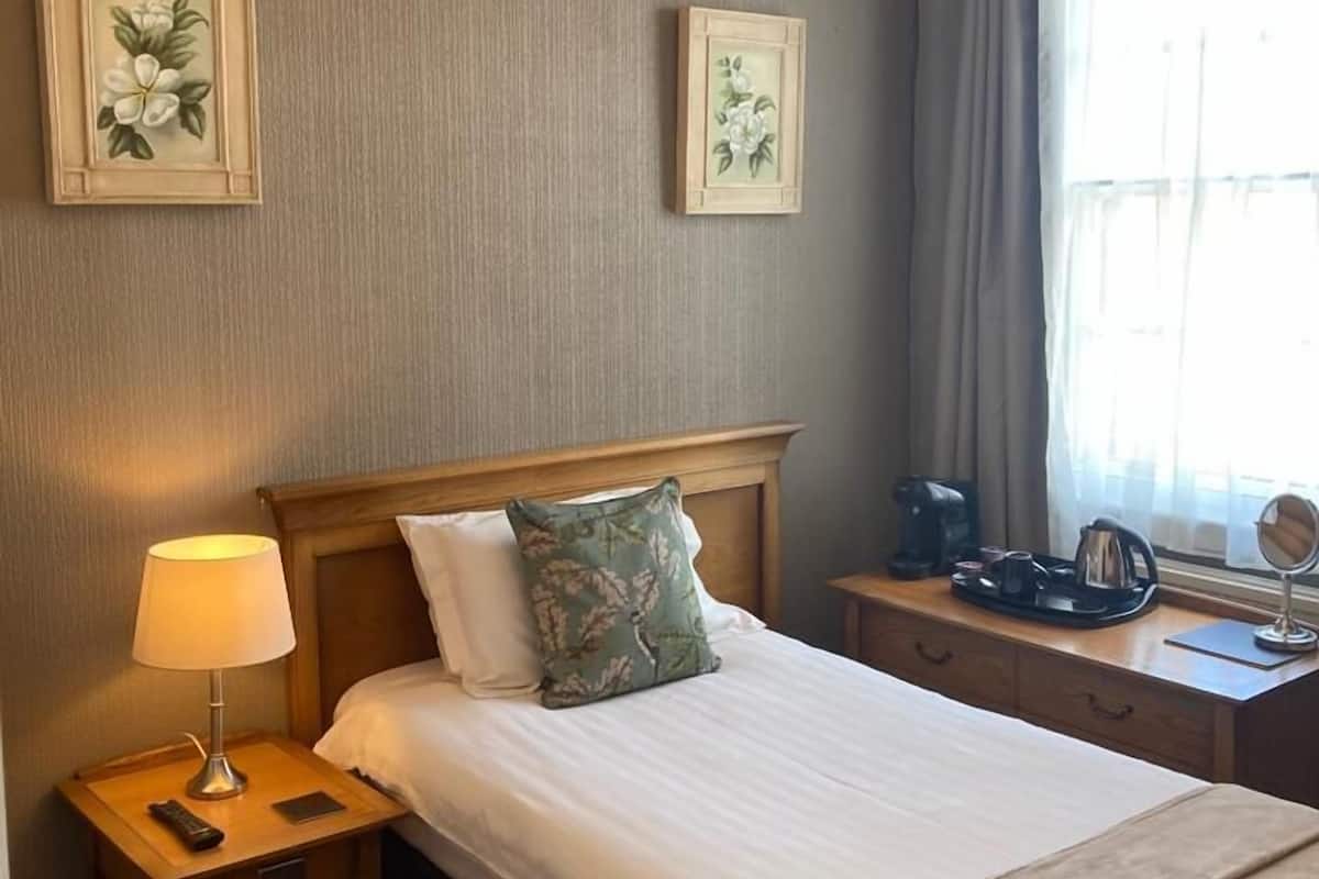 A hotel room with a bed, a nightstand with a lamp, a bedside table with a tray, and a window with curtains.