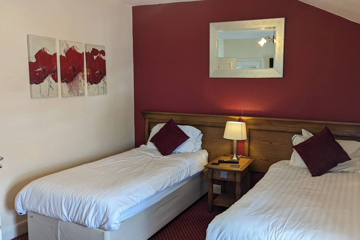 A hotel room with two single beds, a nightstand with a lamp, a mirror, and artwork on the wall.