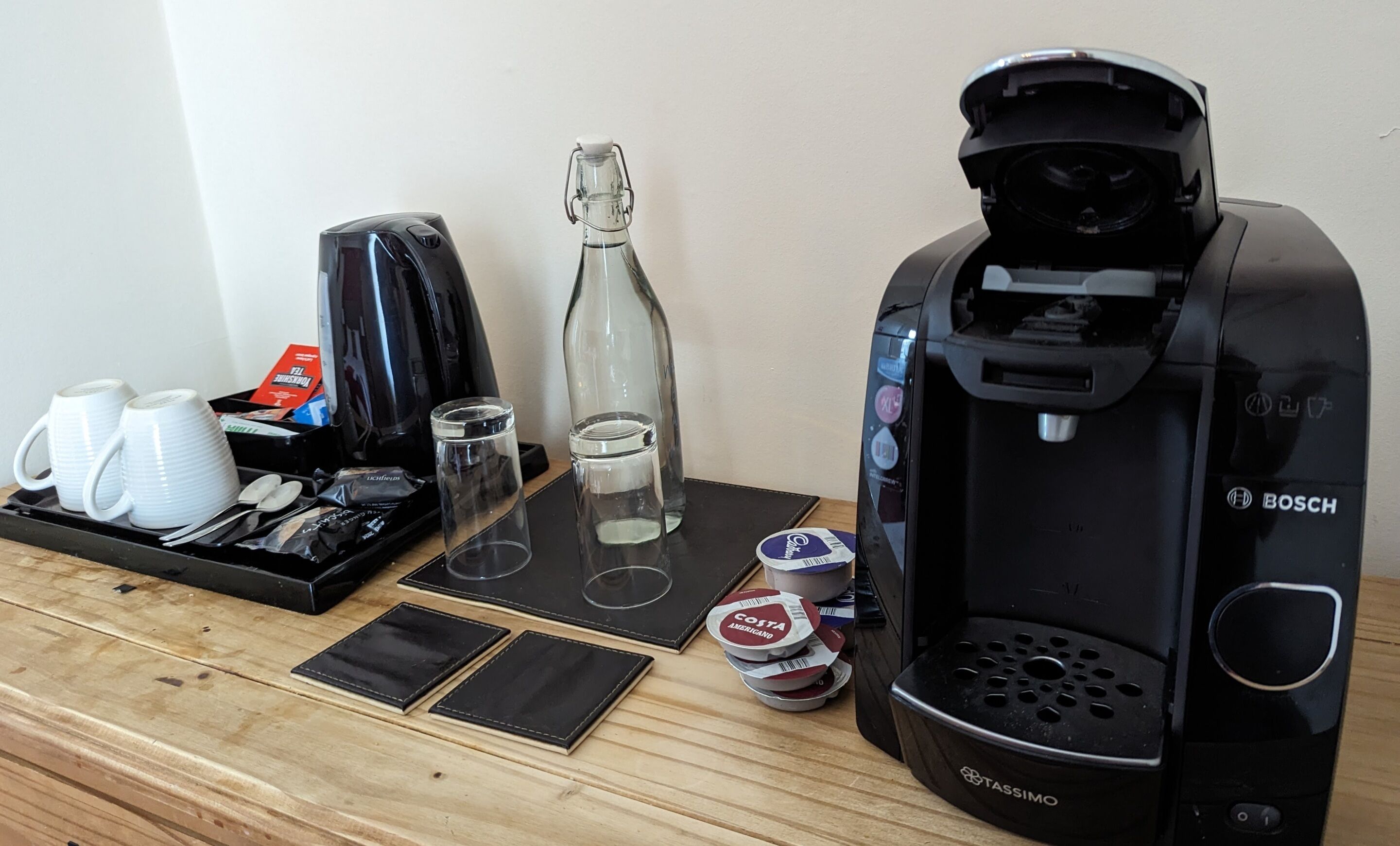 Una cafetera, dos tazas, un vaso, una botella y una bandeja con utensilios y un recipiente pequeño sobre una mesa de madera.