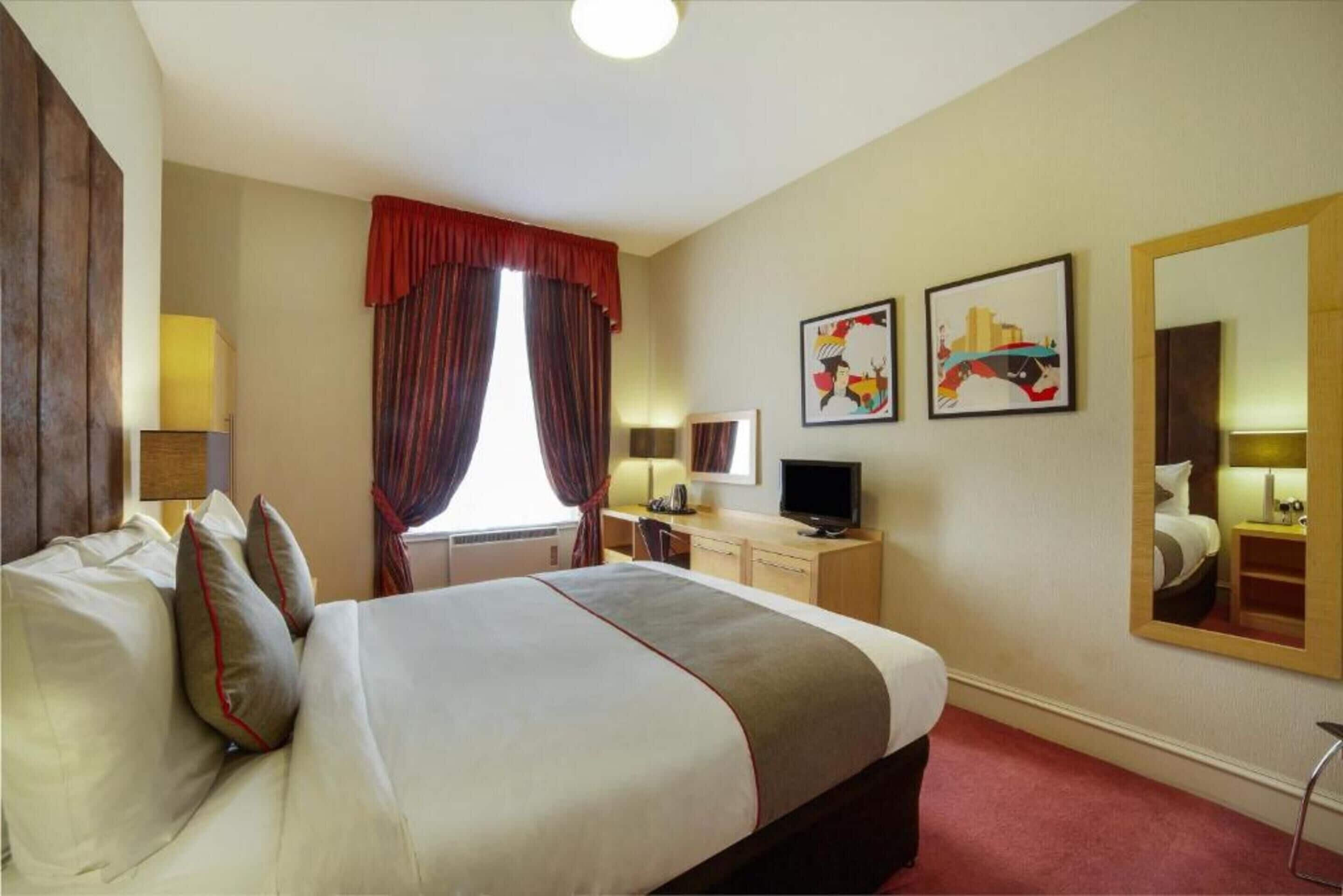 A hotel room with a bed, a desk, a chair, a TV, and artwork on the walls.