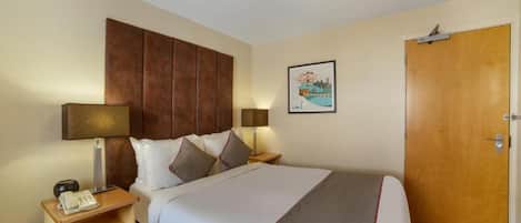 Habitación de hotel con cama, mesitas de noche, teléfono, lámpara, puerta y un cuadro colgado en la pared.
