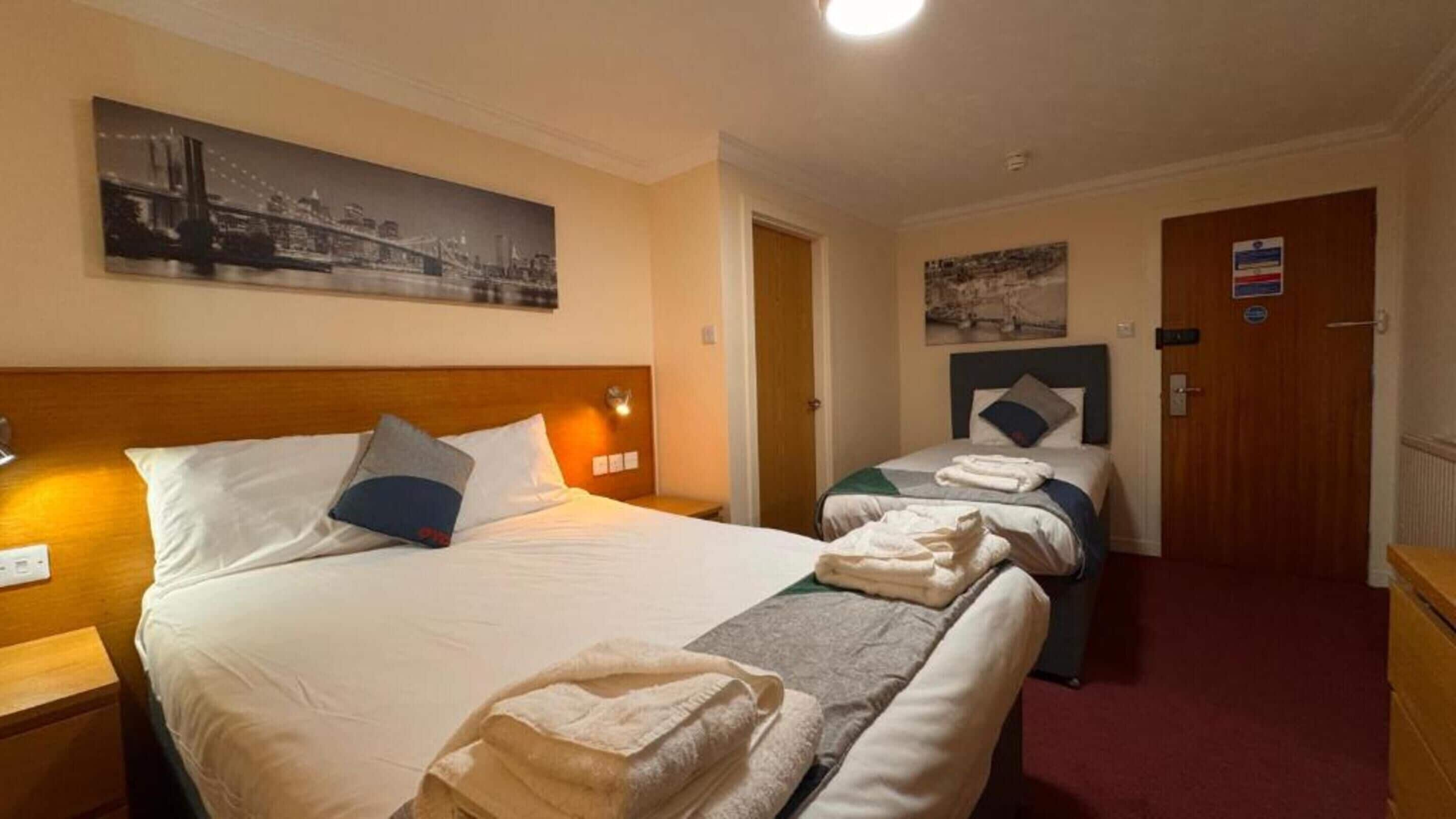A hotel room with two beds, a large bed on the left and a smaller bed on the right. There is a painting on the wall above the large bed, a door on the right, and a small table with a lamp on the left.