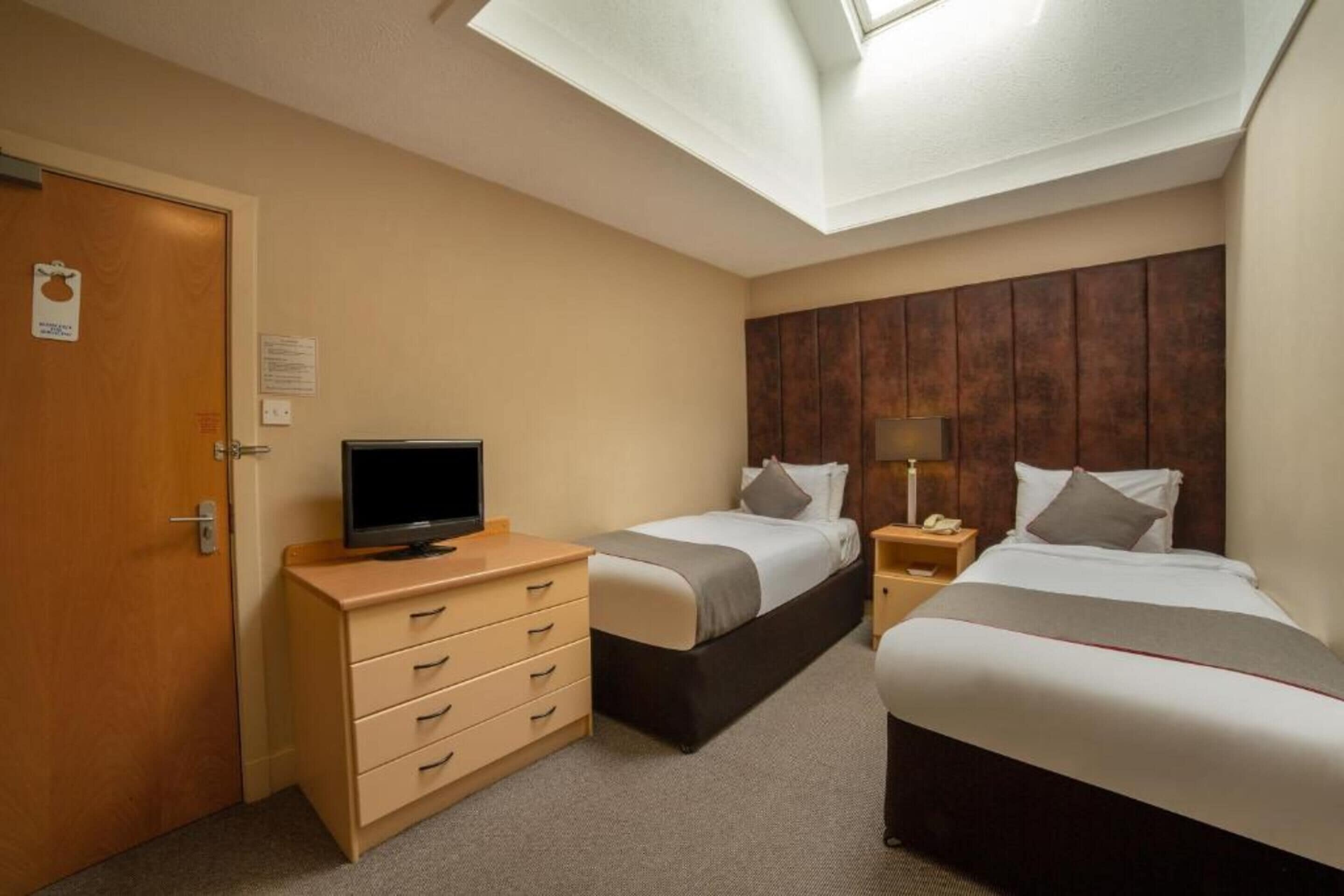 A hotel room with two beds, a TV on a dresser, a wooden headboard, and a skylight.