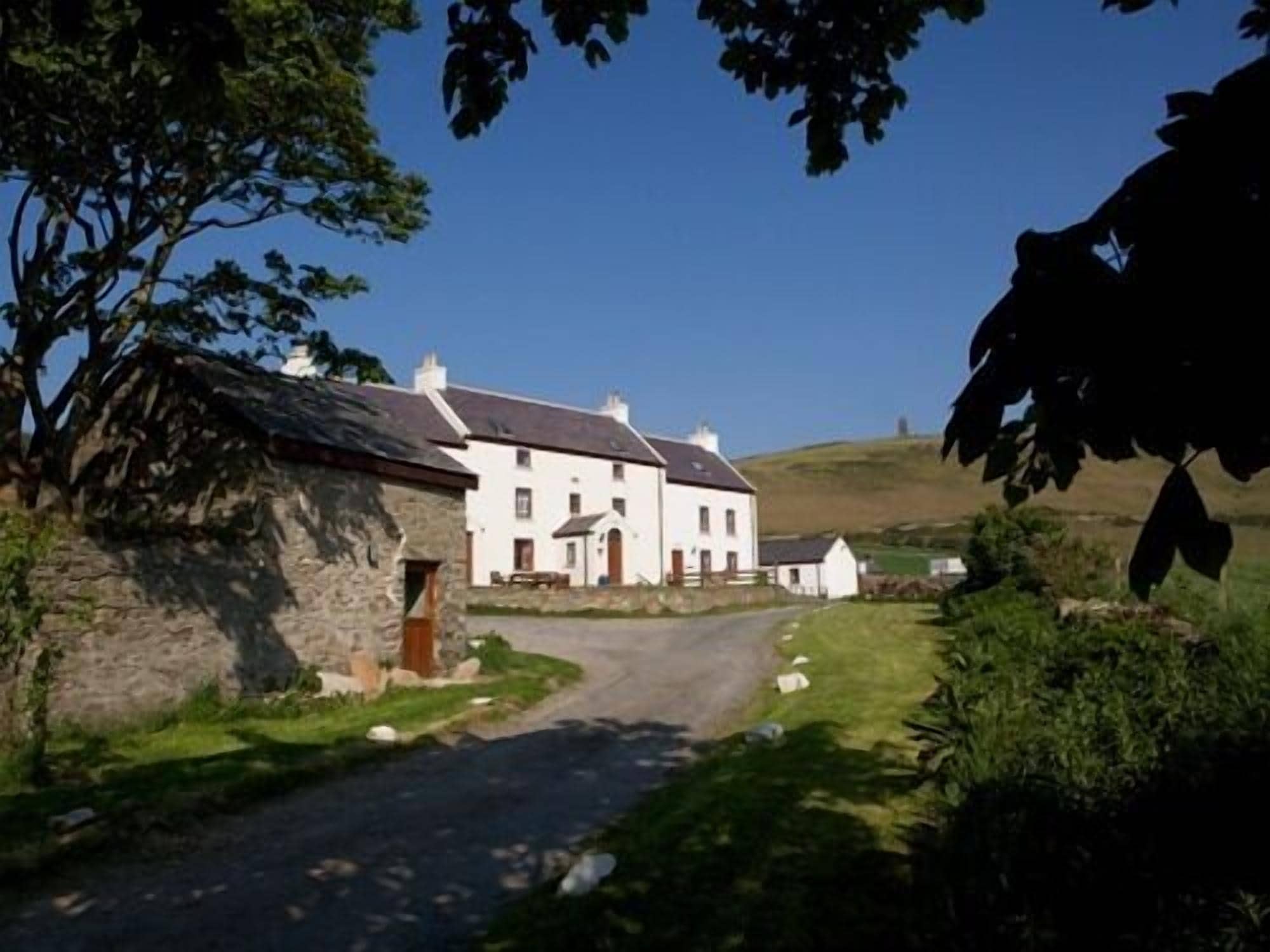 A white building with a stone structure to the left, a paved road, and a grassy area with a hill in the background.