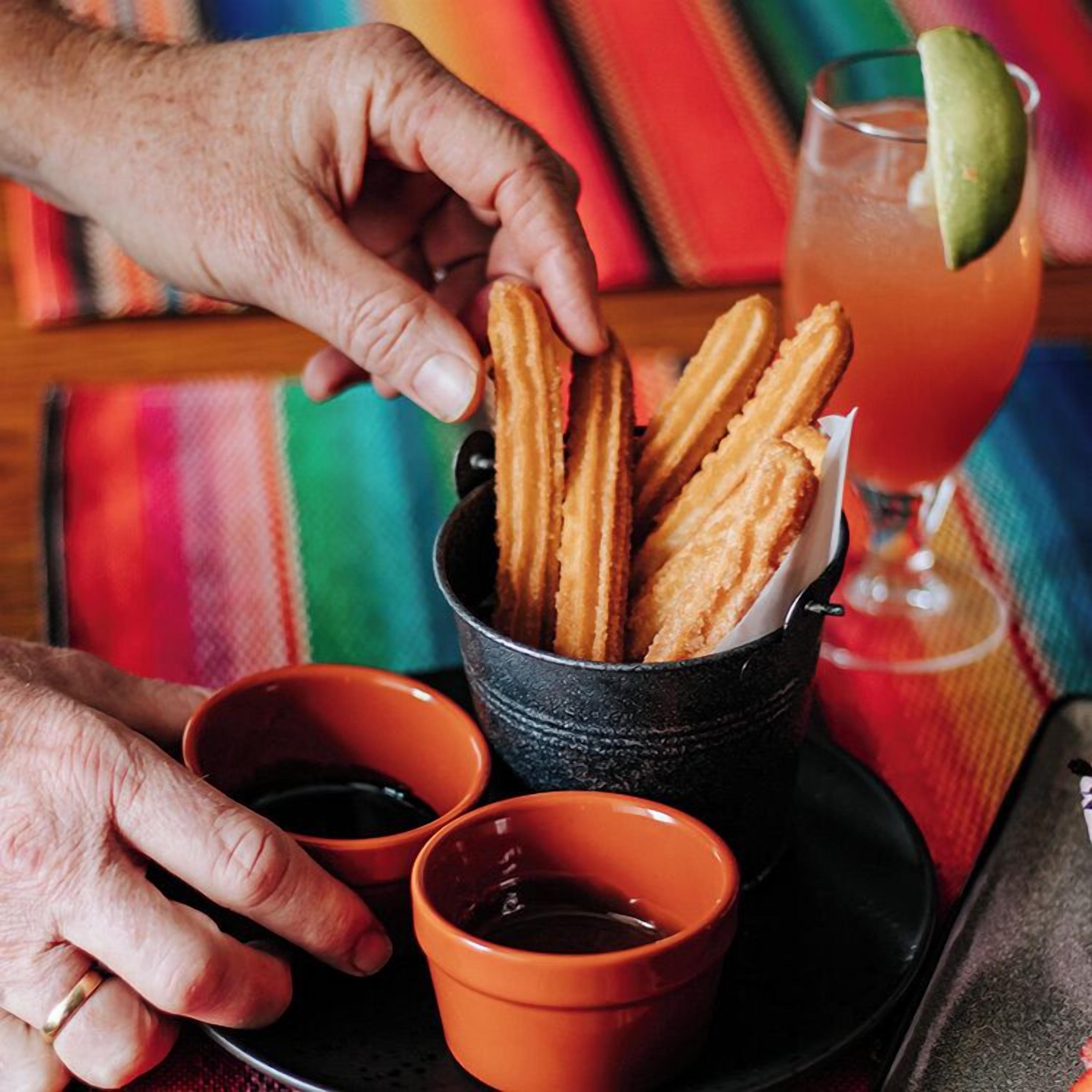 A person holding churros, with dipping sauces and a drink in the background.