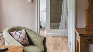 A room with a wooden table, a green armchair, a wooden cabinet, and a doorway leading to a bathroom with a shower and a towel hanging on the wall. - Northover Manor Hotel (Yeovil)