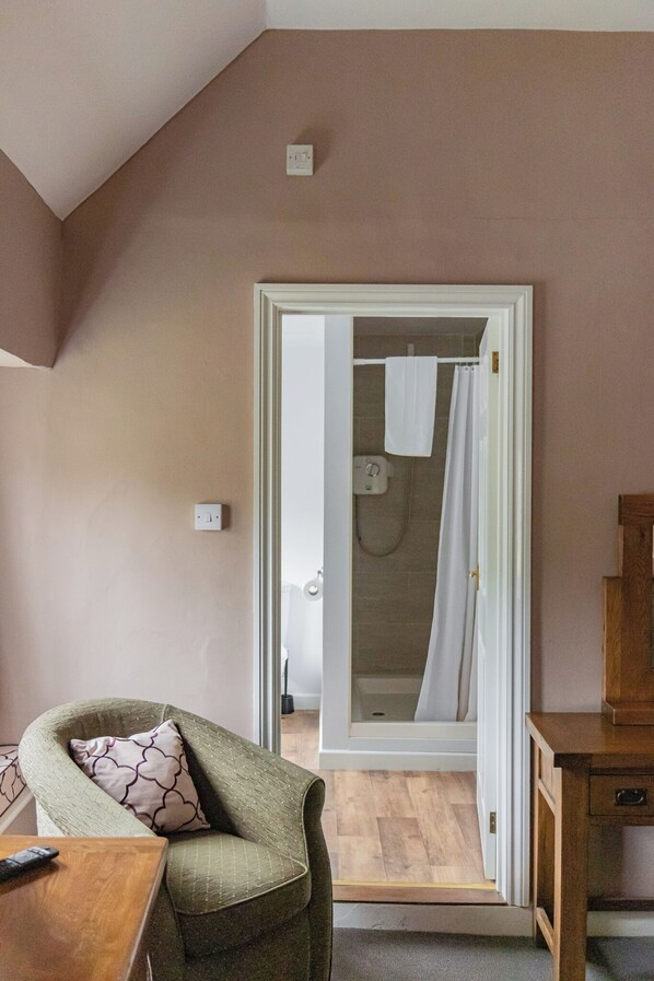 A room with a wooden table, a green armchair, a wooden cabinet, and a doorway leading to a bathroom with a shower and a towel hanging on the wall. - Northover Manor Hotel (Yeovil)