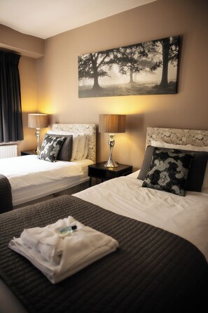 A hotel room with two beds, each with a headboard, a bedside table with a lamp, and a large painting on the wall. - Northover Manor Hotel (Yeovil)