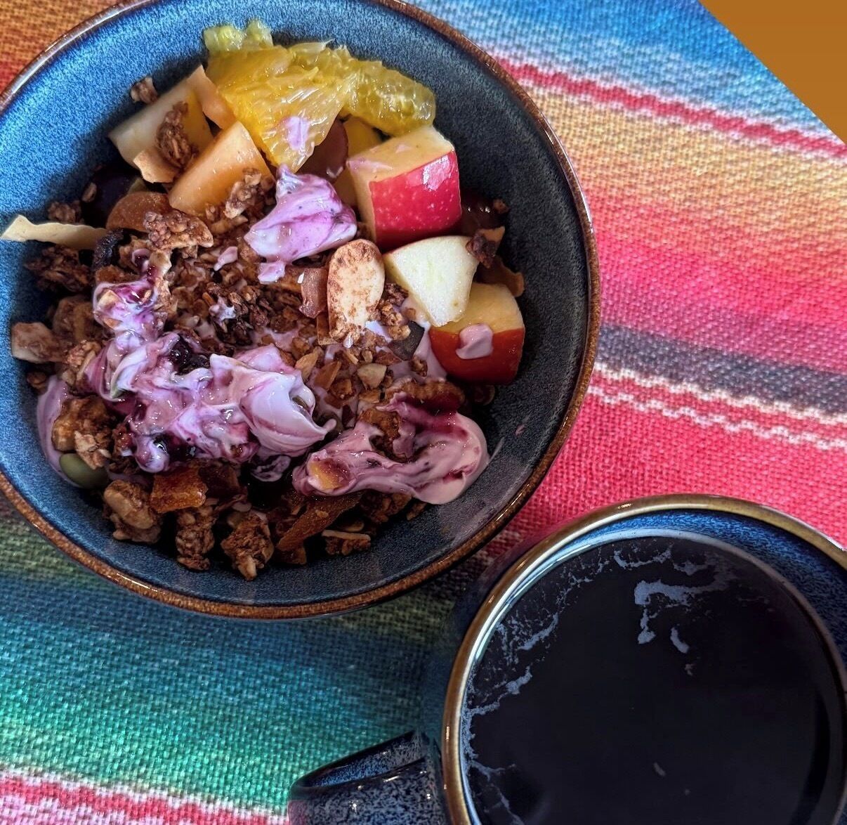 A bowl of granola with fruits and a cup of coffee on a colorful fabric.