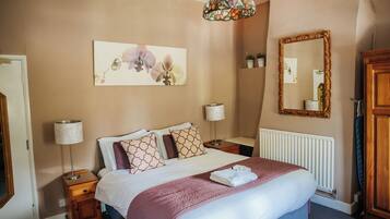 Un dormitorio con cama, lámparas de noche, un espejo, radiador y una obra de arte floral en la pared.