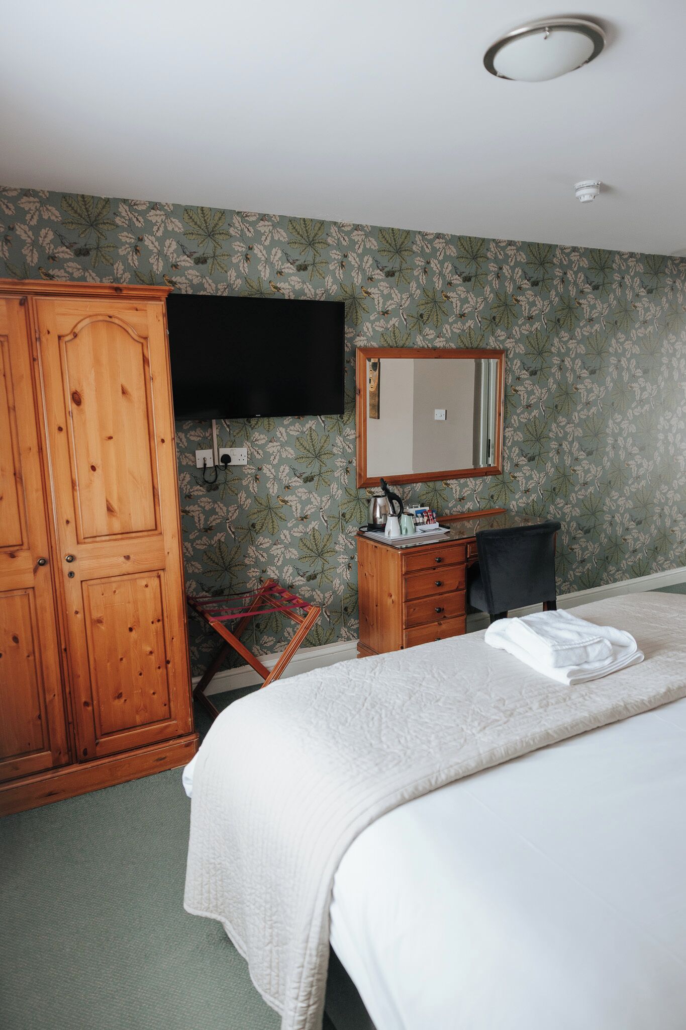 A hotel room with a bed, a wooden wardrobe, a desk with a chair, a flat-screen TV, and a mirror.