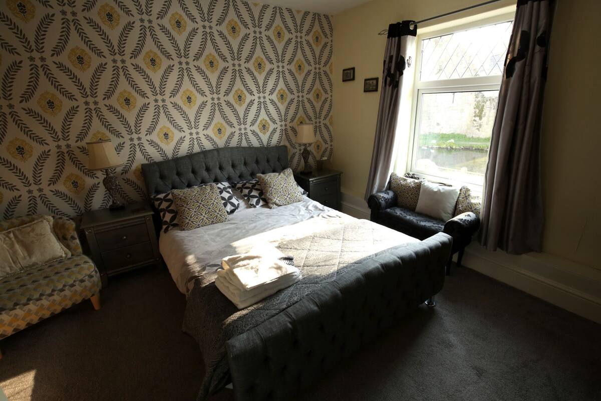 A bedroom with a large bed, a nightstand, a chair, and a window with curtains.