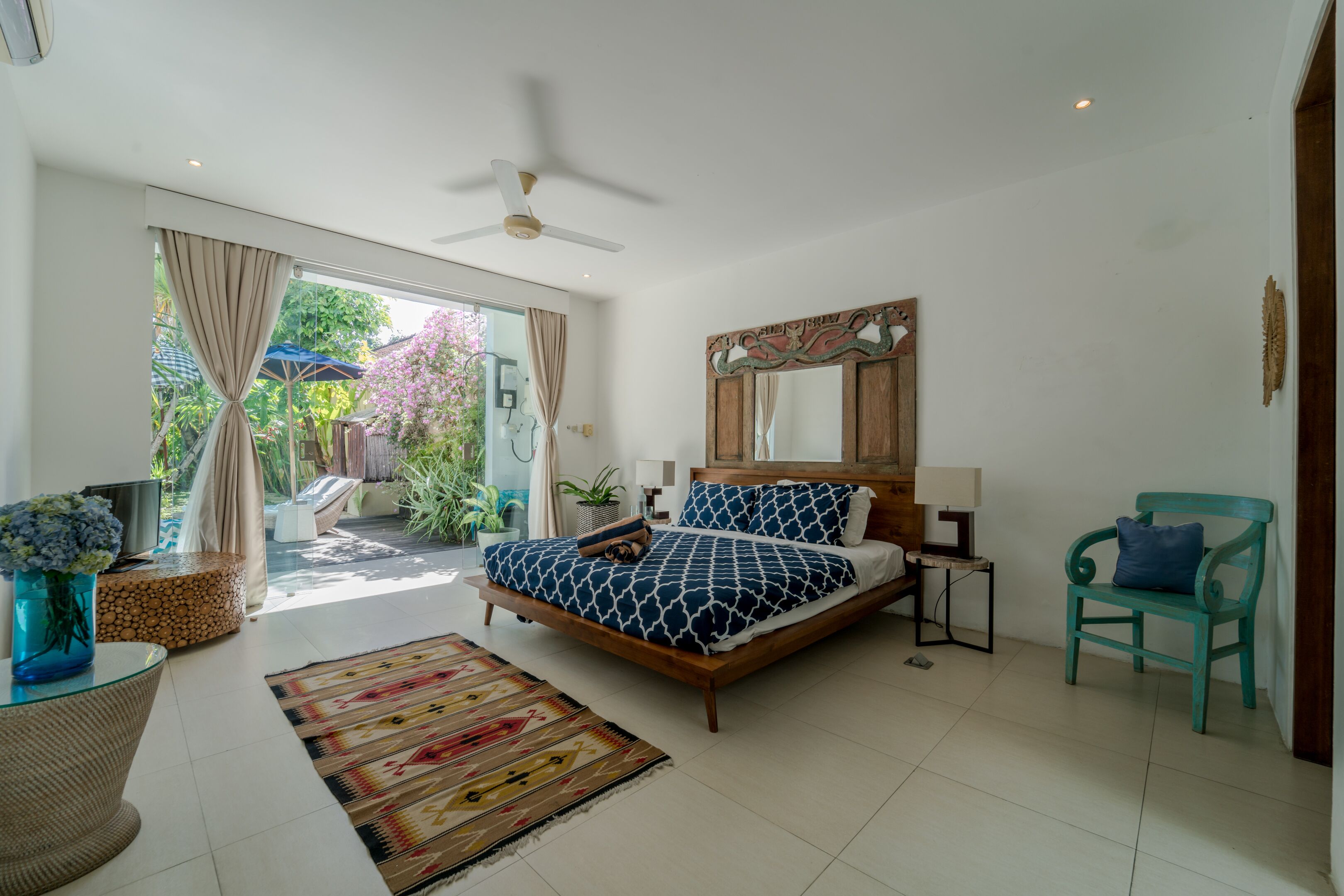 A bedroom with a bed, a ceiling fan, a wooden headboard, a mirror, a chair, a small table with a lamp, and a view of the outdoors.