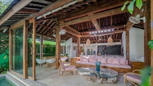 A modern outdoor living area with a wooden ceiling, glass walls, and a pink sofa. - Villa Saffira (Seminyak)