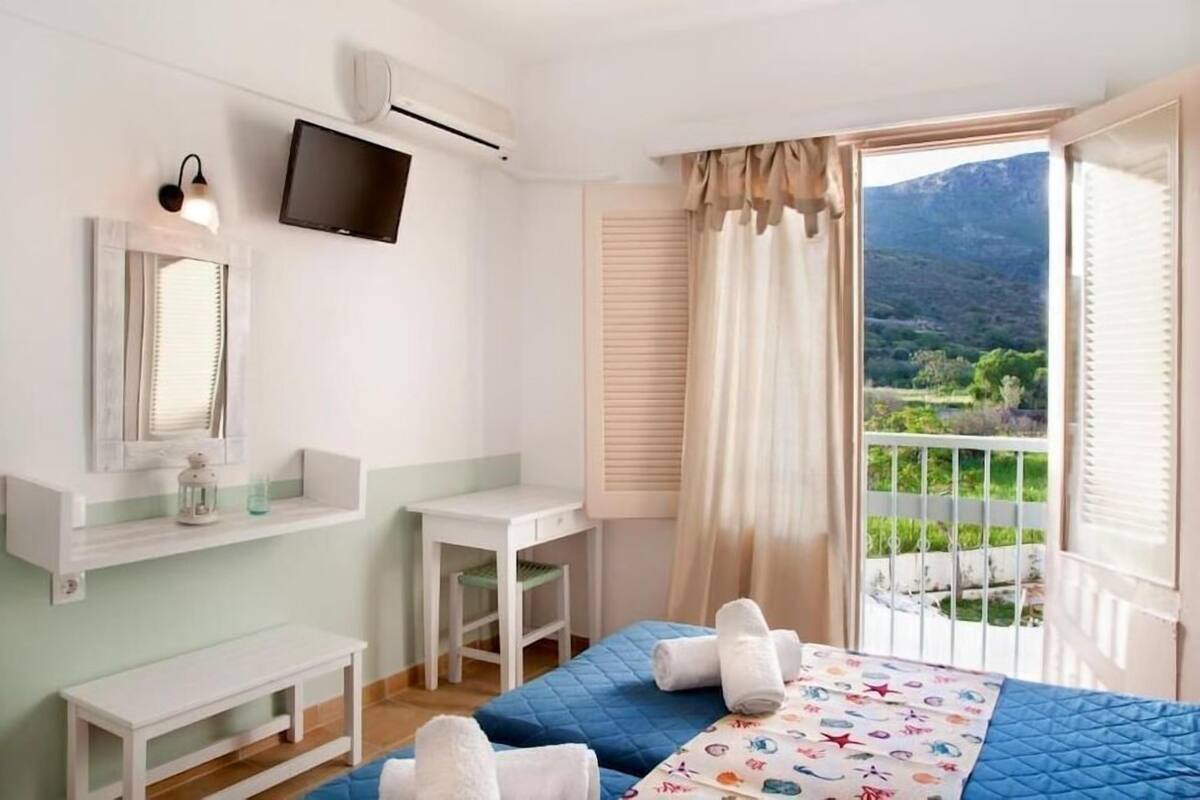 A bedroom with a bed, a desk, a chair, a TV, and a balcony with a mountain view.